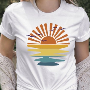 May include: White t-shirt with a retro sunset graphic featuring a sun, ocean waves, and birds flying in the sky.