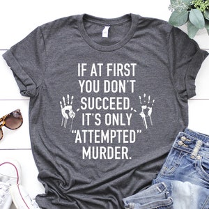 May include: Dark gray t-shirt with the text "IF AT FIRST YOU DON'T SUCCEED, IT'S ONLY 'ATTEMPTED' MURDER." The shirt is paired with blue jeans, white sneakers, and sunglasses. The shirt has a crew neck and short sleeves.