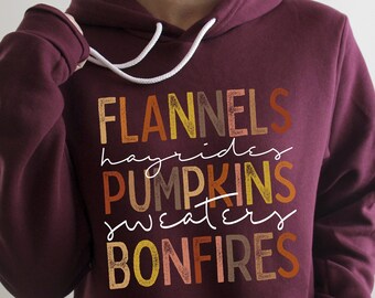 May include: Burgundy hoodie with a white graphic print that says "Flannels, Hayrides, Pumpkins, Sweaters, Bonfires". The text is in a variety of autumn colours.
