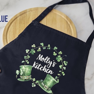 May include: Navy blue apron with a St. Patrick's Day theme. The apron features a green shamrock wreath design with two green top hats and the text "Molly's Kitchen" in white. A wooden spoon and cutting board are in the background.