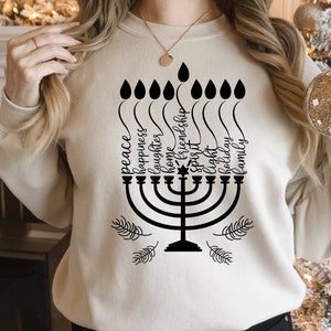 May include: A white sweatshirt with a black menorah design and the words "peace, happiness, laughter, home, friendship, spirit, light, holiday, family" written around it.
