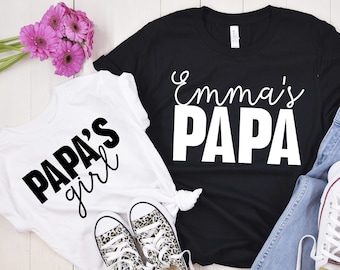 Könnte beinhalten: Zwei weiße T-Shirts mit schwarzem Text. Ein Shirt sagt "Papa's girl" und das andere sagt "Emma's Papa".