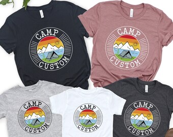 May include: Four t-shirts with a camping theme. The shirts are in different colors, including black, gray, white, and dark gray. Each shirt features a circular design with the text "Camp Custom" and a graphic of a tent, mountains, and a sun.