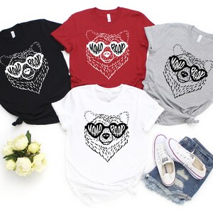 May include: Four t-shirts with a black and white illustration of a bear wearing heart-shaped sunglasses with the text "WILD ROAR" inside the sunglasses. The shirts are in black, red, grey, and white.