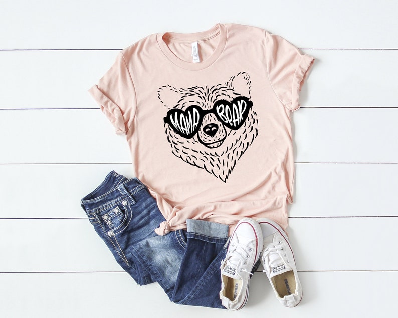 May include: A light pink t-shirt with a black and white illustration of a bear wearing sunglasses that say "Mama Bear". The shirt is laying on a white wooden surface with a pair of blue jeans and white sneakers.