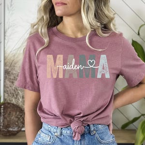 May include: A mauve t-shirt with the word "MAMA" in colourful block letters and the name "aiden" written in cursive with a heart above it.