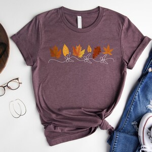 May include: A maroon t-shirt featuring a graphic of autumn leaves in shades of orange, yellow, and brown. The leaves are arranged in a row and have a simple, minimalist design.