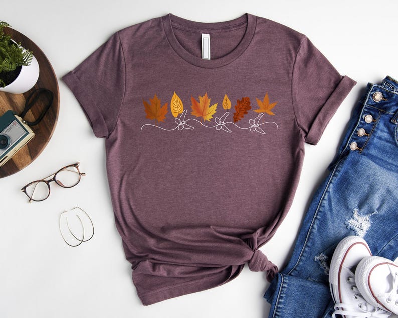 May include: A maroon t-shirt featuring a graphic of autumn leaves in shades of orange, yellow, and brown. The leaves are arranged in a row and have a simple, minimalist design.