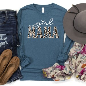 May include: A teal blue long-sleeved shirt with the text "girl mama" in white with a leopard print pattern. The shirt is laid out on a white surface with a pair of blue jeans, brown shoes, a floral scarf, and a brown hat.