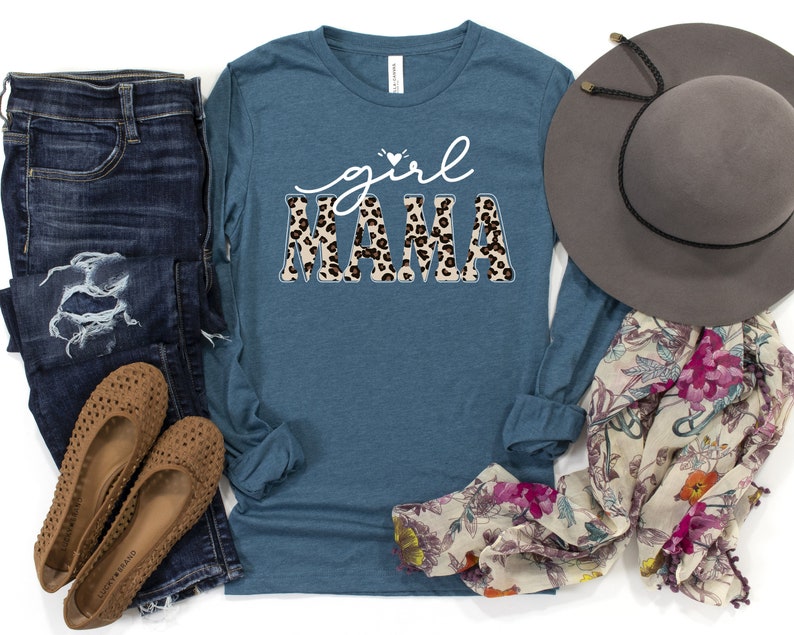 May include: A teal blue long-sleeved shirt with the text "girl mama" in white with a leopard print pattern. The shirt is laid out on a white surface with a pair of blue jeans, brown shoes, a floral scarf, and a brown hat.
