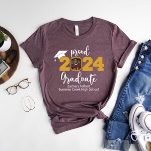 Peut inclure: Un t-shirt marron avec le texte "proud 2024 graduate" en blanc. Le nombre 2024 est en or avec un bonnet de graduation au-dessus. Une photo est dans le nombre 2024. Le texte "Zachary Tolbert Summer Creek High School" est sous le nombre 2024.