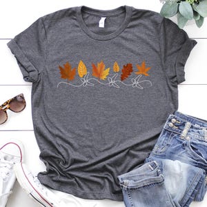 May include: A dark grey t-shirt with a white outline of a row of autumn leaves in shades of orange, yellow, and brown.