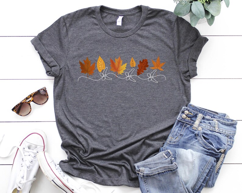 May include: A dark grey t-shirt with a white outline of a row of autumn leaves in shades of orange, yellow, and brown.
