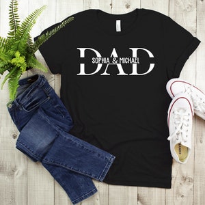 Puede incluir: Camiseta negra con texto blanco que dice "DAD" con los nombres "Sophia & Michael" debajo. La camiseta est&aacute; colocada sobre una superficie de madera con un par de zapatillas blancas y unos vaqueros azules.