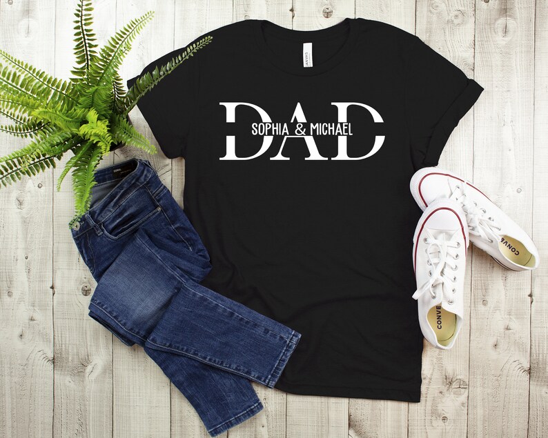 Puede incluir: Camiseta negra con texto blanco que dice "DAD" con los nombres "Sophia & Michael" debajo. La camiseta est&aacute; colocada sobre una superficie de madera con un par de zapatillas blancas y unos vaqueros azules.