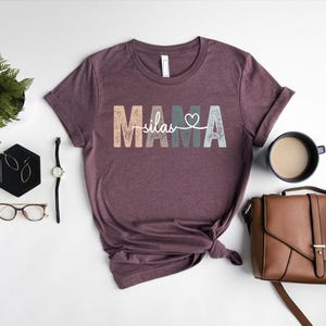 May include: A maroon t-shirt with the text "MAMA" in colourful letters with a heart and the word "silas" in white text.