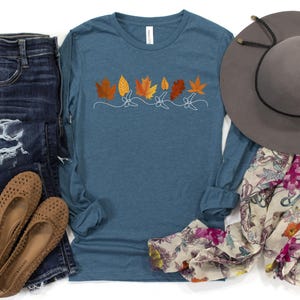 May include: A long-sleeved, teal blue t-shirt with a row of orange and yellow leaf designs. The shirt is on a white background with a pair of blue jeans, a brown hat, and a floral scarf.