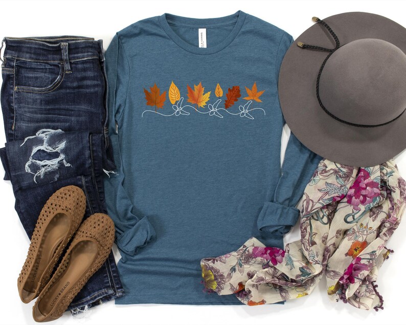 May include: A long-sleeved, teal blue t-shirt with a row of orange and yellow leaf designs. The shirt is on a white background with a pair of blue jeans, a brown hat, and a floral scarf.