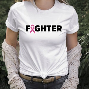 Pu&ograve; includere: Maglietta bianca con un nastro rosa e la parola "FIGHTER" stampata in nero.