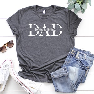 Puede incluir: Una camiseta gris oscuro con la palabra "DAD" en letras blancas. La palabra "DAD" est&aacute; separada por una l&iacute;nea horizontal con el texto "Sophia & Michael" escrito encima de la l&iacute;nea.