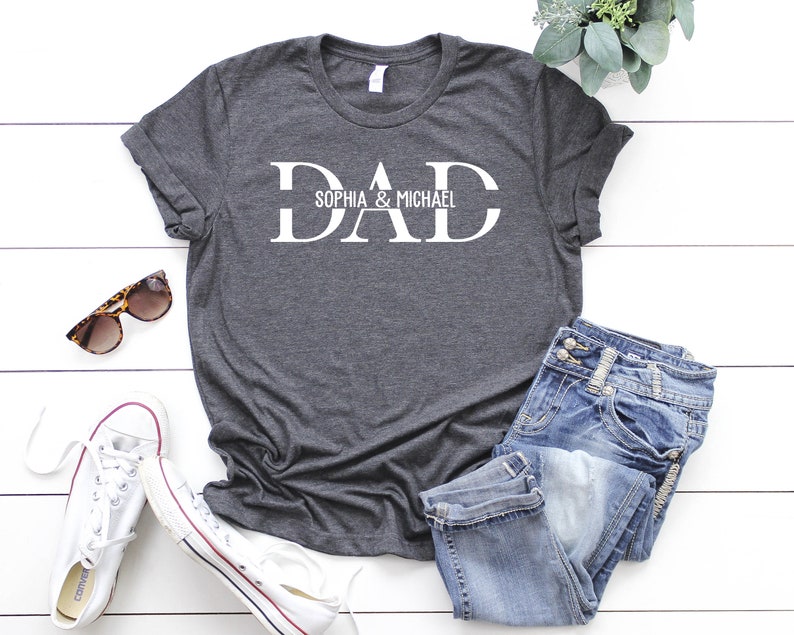 Puede incluir: Una camiseta gris oscuro con la palabra "DAD" en letras blancas. La palabra "DAD" est&aacute; separada por una l&iacute;nea horizontal con el texto "Sophia & Michael" escrito encima de la l&iacute;nea.