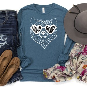 May include: A teal long-sleeve shirt with a white bear wearing sunglasses that say "Mama Bear". The bear is wearing sunglasses with the text "Mama Bear" on them. The shirt is laid out on a white background with a pair of brown flats, blue jeans, a floral scarf, and a gray hat.
