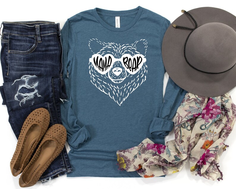 May include: A teal long-sleeve shirt with a white bear wearing sunglasses that say "Mama Bear". The bear is wearing sunglasses with the text "Mama Bear" on them. The shirt is laid out on a white background with a pair of brown flats, blue jeans, a floral scarf, and a gray hat.