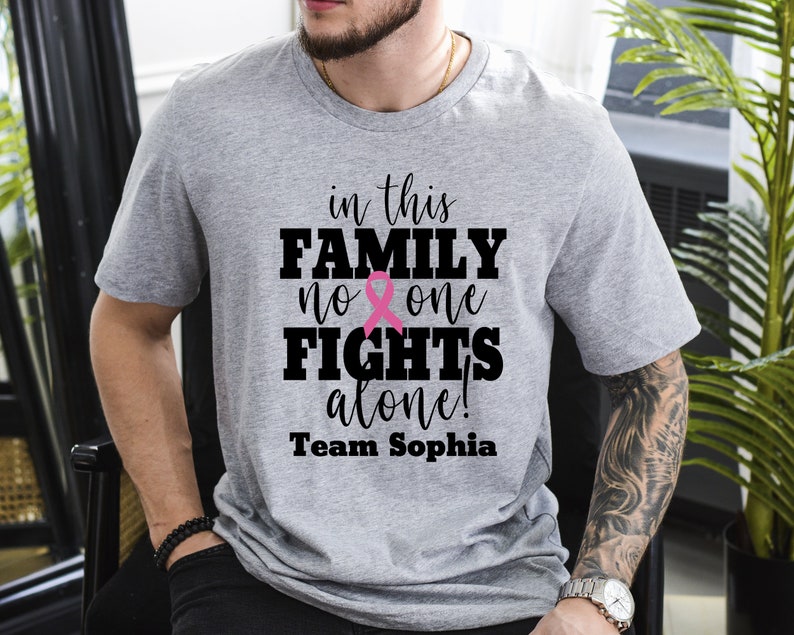 Pu&ograve; includere: Maglietta grigia con il testo "in this FAMILY no one FIGHTS alone! Team Sophia" e un grafico a nastro rosa.