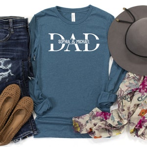 Puede incluir: Una camiseta de manga larga azul verdosa con la palabra "DAD" en letras blancas en bloque y el texto "Sophia & Michael" debajo. La camiseta est&aacute; colocada sobre una superficie blanca con un par de zapatos marrones, unos vaqueros azules, una bufanda floral y un sombrero gris.