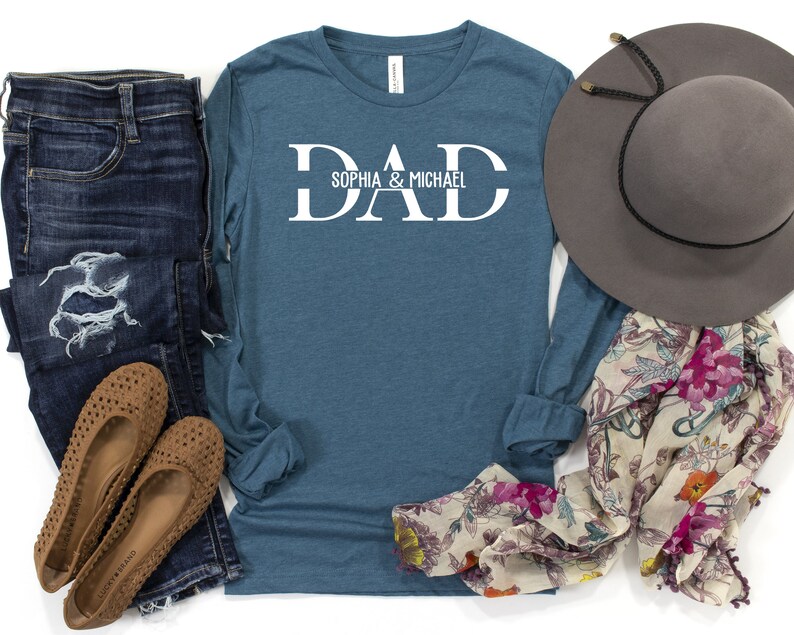 Puede incluir: Una camiseta de manga larga azul verdosa con la palabra "DAD" en letras blancas en bloque y el texto "Sophia & Michael" debajo. La camiseta est&aacute; colocada sobre una superficie blanca con un par de zapatos marrones, unos vaqueros azules, una bufanda floral y un sombrero gris.