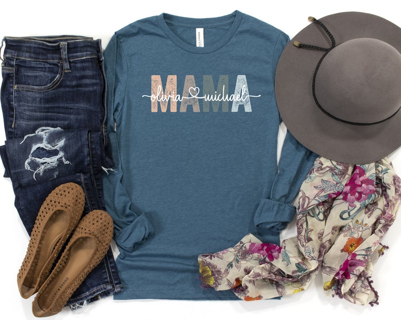 May include: A teal long-sleeved shirt with the word "MAMA" in colourful letters and the text "olivia & michael" underneath. The shirt is paired with blue jeans, brown flats, a floral scarf, and a brown hat.