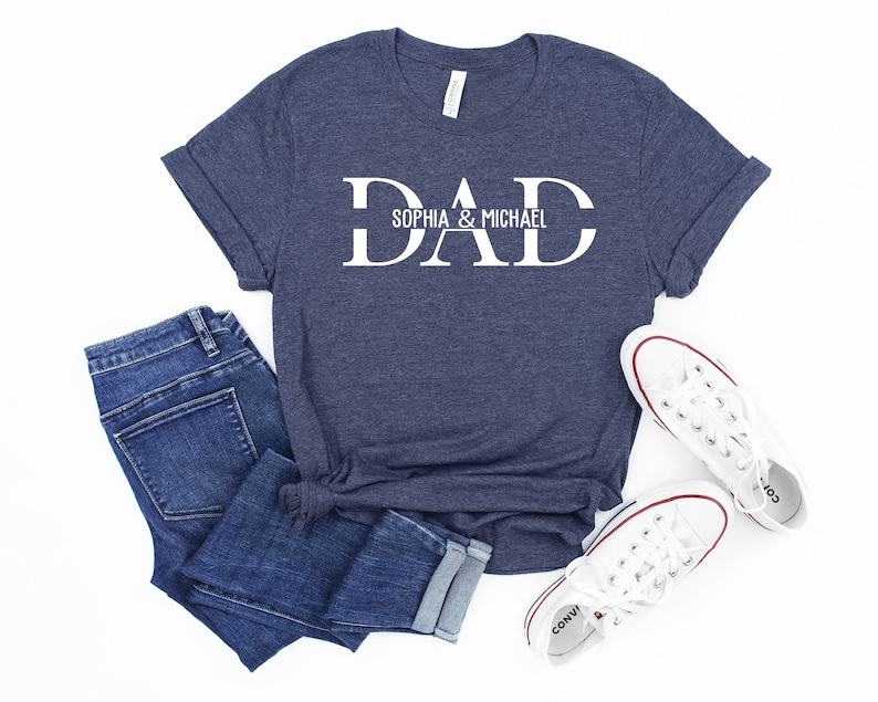 Puede incluir: Una camiseta azul marino con texto blanco que dice "DAD" con los nombres "Sophia & Michael" debajo. La camiseta est&aacute; sobre una superficie blanca con un par de zapatillas blancas y unos vaqueros azules.