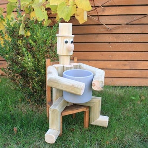 Könnte beinhalten: Holzfigur eines Menschen, der auf einem Stuhl sitzt und einen grauen Blumentopf hält. Die Figur hat einen Hut, Augen und eine Nase.