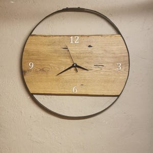 Könnte beinhalten: Eine runde Wanduhr mit einem hellbraunen Holzgesicht und einem dunklen Metallrahmen. Das Zifferblatt hat weiße Ziffern an den Positionen 3, 6, 9 und 12 Uhr, mit schwarzen Zeigern. Die Uhr ist an einer beigen Wand befestigt.