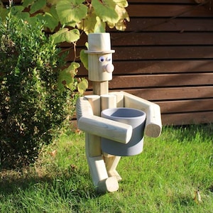 Könnte beinhalten: Holzfigur eines Menschen mit Hut, der einen grauen Blumentopf hält. Die Figur steht auf einem grünen Rasen.