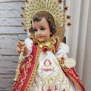 Peut inclure: Poupée détaillée d'une figure religieuse, ornée d'un halo doré serti de pierres rouges. La poupée porte une robe brodée blanche et rouge avec des accents dorés, tenant un bâton et une croix dorés. La poupée est assise sur une petite chaise.