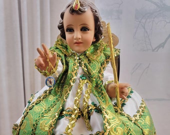 Puede incluir: Una figurita de porcelana de un niño vestido con un vestido blanco con una capa bordada en verde y dorado. La figurita sostiene un cetro dorado y un pequeño objeto redondo. La figurita está sentada en una silla de madera. El texto "Mexshop Boutique" es visible en la imagen.