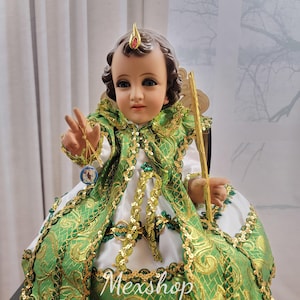 Peut inclure: Une figurine en porcelaine d'un enfant vêtu d'une robe blanche avec une cape brodée de vert et d'or. La figurine tient un sceptre doré et un petit objet rond. La figurine est assise sur une chaise en bois. Le texte "Mexshop Boutique" est visible sur l'image.