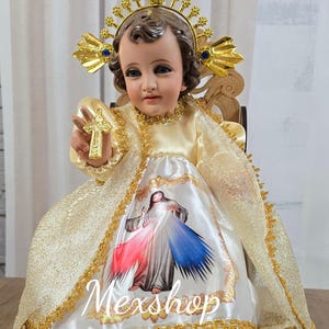 May include: A religious figurine of a child, wearing a gold crown with blue accents and holding a gold cross. The figurine is dressed in a cream-colored gown with a printed image and the text "Mexshop Boutique".