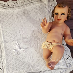 Op de afbeelding: Wit gebreide babydeken met een delicate kantzoom en een geborduurd ontwerp van een baby Jezus. De deken ligt op een houten oppervlak.
