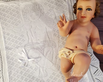 Könnte beinhalten: Weiße gestrickte Babydecke mit zarter Spitzenbordüre und einem gestickten Motiv eines Jesuskindes. Die Decke liegt auf einer Holzoberfläche.