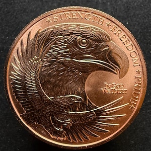 May include: A copper-colored coin featuring a detailed eagle design. The coin's edge is inscribed with the words "STRENGTH FREEDOM PRIDE" and the phrase "IN GOD WE TRUST". The eagle is depicted with intricate feather details.