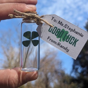 May include: A clear glass vial containing a single four-leaf clover, sealed with a cork stopper and tied with twine. A white rectangular paper tag is attached, featuring the printed text: To: Ms Stephanie, GOOD LUCK, From: Randy.