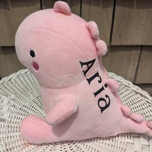 Puede incluir: Un dinosaurio de peluche rosa con el nombre "Aria" bordado en negro en el lateral.