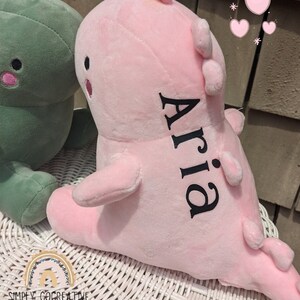 Puede incluir: Juguete de dinosaurio de peluche rosa con el nombre "Aria" bordado en la espalda. El dinosaurio est&aacute; sentado sobre una superficie de mimbre blanco.