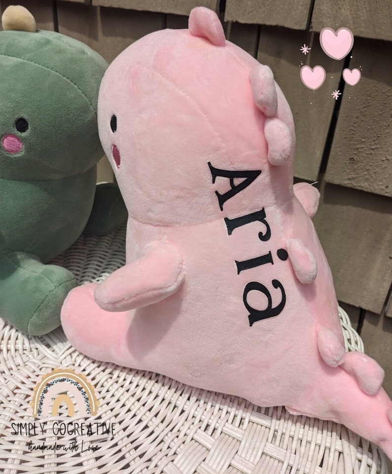 Puede incluir: Juguete de dinosaurio de peluche rosa con el nombre "Aria" bordado en la espalda. El dinosaurio est&aacute; sentado sobre una superficie de mimbre blanco.