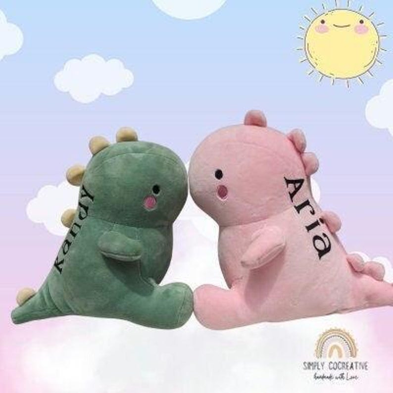 Puede incluir: Dos peluches de dinosaurio, uno verde y otro rosa, con los nombres bordados "Kendy" y "Aria".