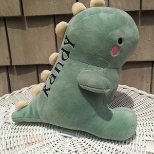 Puede incluir: Juguete de dinosaurio de peluche verde con p&uacute;as amarillas y una mejilla rosa. El dinosaurio tiene el nombre "Randy" bordado en un lado.