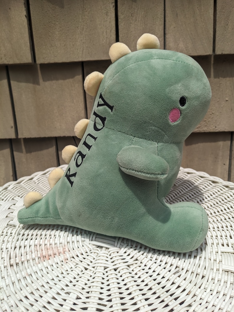 Puede incluir: Juguete de dinosaurio de peluche verde con p&uacute;as amarillas y una mejilla rosa. El dinosaurio tiene el nombre "Randy" bordado en un lado.