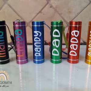 May include: Six personalised mini torches in different colours, including black, pink, blue, green, red, and gold. Each torch has a name printed on it: mama, linda, randy, dad, dada, and marilyn.